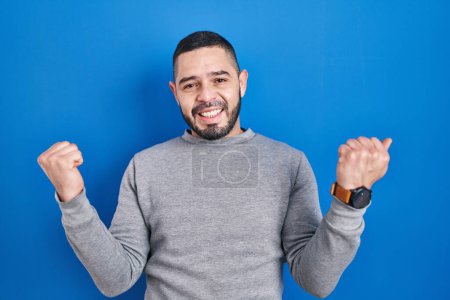 Photo for Hispanic man standing over blue background celebrating surprised and amazed for success with arms raised and eyes closed. winner concept. - Royalty Free Image