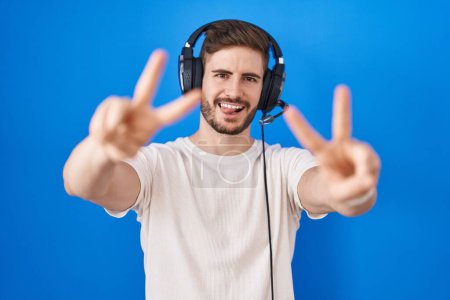 Téléchargez les photos : Homme hispanique avec barbe écoutant de la musique portant des écouteurs souriant avec la langue montrant les doigts des deux mains faisant signe de victoire. numéro deux. - en image libre de droit