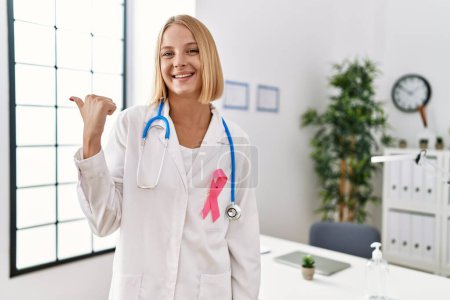 Téléchargez les photos : Jeune médecin caucasien femme portant un ruban rose cancer pointant le pouce vers le côté souriant heureux avec la bouche ouverte - en image libre de droit