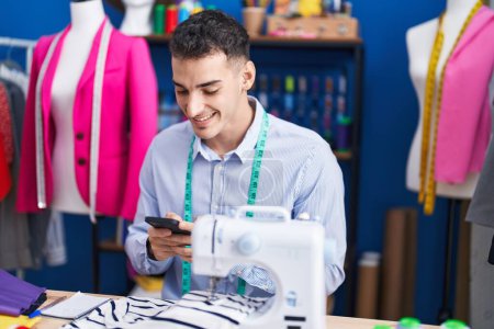 Téléchargez les photos : Jeune homme hispanique tailleur à l'aide d'un smartphone au studio de couture - en image libre de droit
