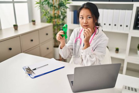 Photo for Young asian doctor woman holding support green ribbon serious face thinking about question with hand on chin, thoughtful about confusing idea - Royalty Free Image
