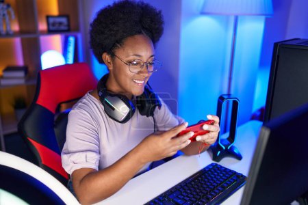 Foto de Streamer mujer afroamericana usando teléfono inteligente jugando videojuego en la sala de juegos - Imagen libre de derechos