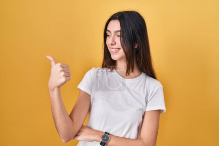 Photo for Young beautiful woman standing over yellow background looking proud, smiling doing thumbs up gesture to the side - Royalty Free Image