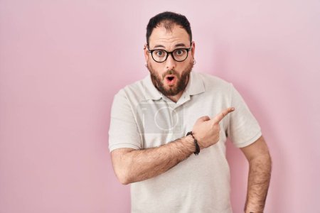 Photo for Plus size hispanic man with beard standing over pink background surprised pointing with finger to the side, open mouth amazed expression. - Royalty Free Image