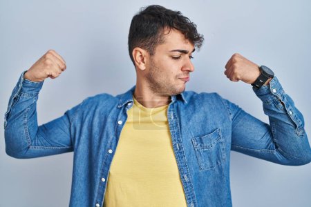 Téléchargez les photos : Jeune homme hispanique debout sur fond bleu montrant les muscles des bras souriant fier. concept de fitness. - en image libre de droit