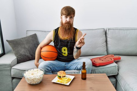 Photo for Caucasian man with long beard holding basketball ball cheering tv game pointing with hand finger to the side showing advertisement, serious and calm face - Royalty Free Image