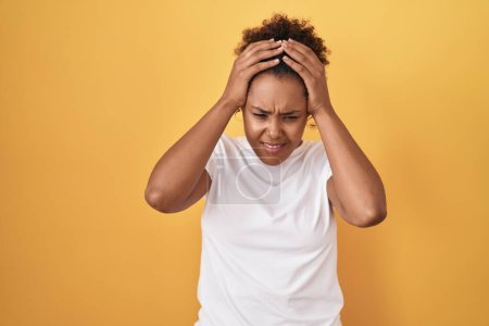 Foto de Young hispanic woman with curly hair standing over yellow background suffering from headache desperate and stressed because pain and migraine. hands on head. - Imagen libre de derechos