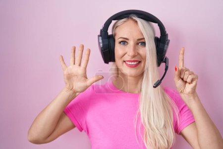 Téléchargez les photos : Femme caucasienne écoutant de la musique à l'aide d'écouteurs montrant et pointant vers le haut avec les doigts numéro six tout en souriant confiant et heureux. - en image libre de droit