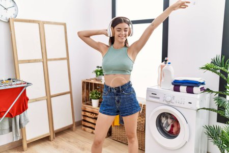 Foto de Young hispanic woman listening to music waiting for washing machine at laundry room - Imagen libre de derechos