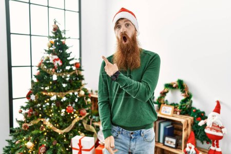 Téléchargez les photos : Homme rousse à longue barbe portant un chapeau de Noël par arbre de Noël surpris pointant du doigt sur le côté, bouche ouverte expression étonnée. - en image libre de droit