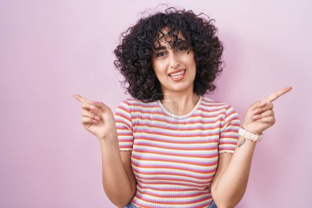 Téléchargez les photos : Jeune femme du Moyen-Orient debout sur fond rose souriant confiant pointant avec les doigts vers différentes directions. espace de copie pour la publicité - en image libre de droit