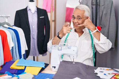 Photo for Middle age man with grey hair dressmaker using sewing machine smiling doing talking on the telephone gesture and pointing to you. call me. - Royalty Free Image