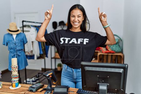 Photo for Young hispanic woman working as staff at retail boutique smiling amazed and surprised and pointing up with fingers and raised arms. - Royalty Free Image