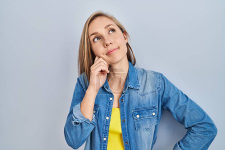 Photo for Young blonde woman standing over blue background with hand on chin thinking about question, pensive expression. smiling with thoughtful face. doubt concept. - Royalty Free Image
