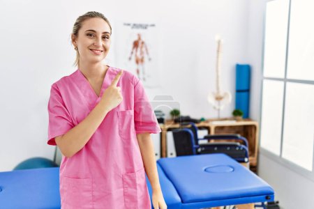 Foto de Young blonde woman working at pain recovery clinic cheerful with a smile of face pointing with hand and finger up to the side with happy and natural expression on face - Imagen libre de derechos