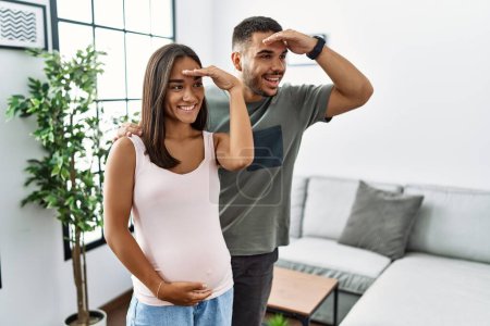 Photo for Young interracial couple expecting a baby, touching pregnant belly very happy and smiling looking far away with hand over head. searching concept. - Royalty Free Image