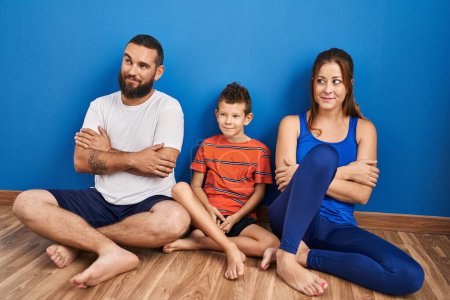 Téléchargez les photos : Famille de trois assis sur le sol à la maison souriant regardant vers le côté et regardant loin en pensant. - en image libre de droit