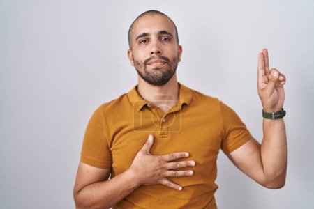 Foto de Hispanic man with beard standing over white background smiling swearing with hand on chest and fingers up, making a loyalty promise oath - Imagen libre de derechos