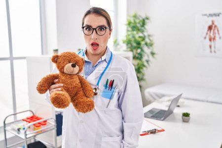 Foto de Young hispanic pediatrician woman holding teddy bear at the clinic scared and amazed with open mouth for surprise, disbelief face - Imagen libre de derechos