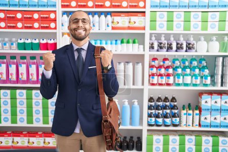 Téléchargez les photos : Homme hispanique avec barbe travaillant comme vendeur à la pharmacie drugstore criant fier, célébrant la victoire et le succès très excité avec les bras levés - en image libre de droit