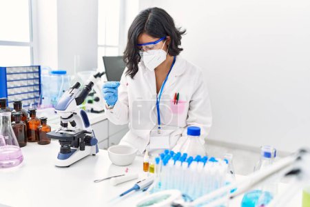 Foto de Mujer latina joven vistiendo uniforme científico y máscara médica sosteniendo píldora en laboratorio - Imagen libre de derechos