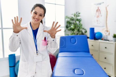 Téléchargez les photos : Jeune médecin femme travaillant à la clinique de récupération de la douleur montrant et pointant vers le haut avec les doigts numéro dix tout en souriant confiant et heureux. - en image libre de droit
