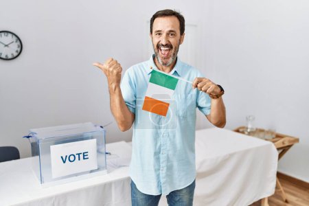 Foto de Hombre de mediana edad con barba en las elecciones de campaña política sosteniendo la bandera de Irlanda señalando el pulgar hacia el lado sonriendo feliz con la boca abierta - Imagen libre de derechos