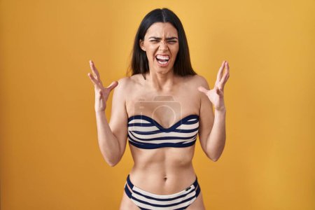 Photo for Young brunette woman wearing bikini over yellow background crazy and mad shouting and yelling with aggressive expression and arms raised. frustration concept. - Royalty Free Image