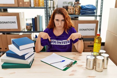 Photo for Young redhead woman wearing volunteer t shirt at donations stand pointing down looking sad and upset, indicating direction with fingers, unhappy and depressed. - Royalty Free Image