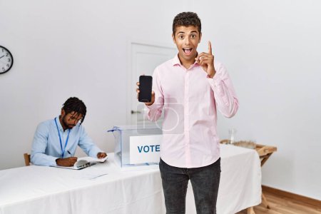Foto de Hombres hispanos jóvenes en las elecciones de campaña política con smartphone sorprendido con una idea o pregunta apuntando con el dedo con la cara feliz, número uno - Imagen libre de derechos