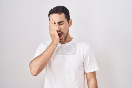 Foto de Handsome hispanic man standing over white background yawning tired covering half face, eye and mouth with hand. face hurts in pain. - Imagen libre de derechos