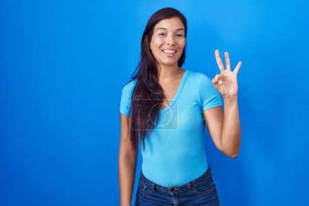 Photo for Young hispanic woman standing over blue background showing and pointing up with fingers number three while smiling confident and happy. - Royalty Free Image