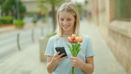 Photo for Young blonde woman using smartphone holding bouquet of flowers at street - Royalty Free Image