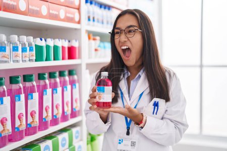 Photo for Young hispanic woman working at pharmacy drugstore holding syrup angry and mad screaming frustrated and furious, shouting with anger. rage and aggressive concept. - Royalty Free Image