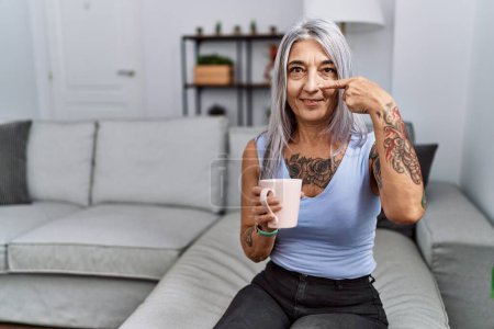 Téléchargez les photos : Femme aux cheveux gris d'âge moyen buvant du café assis sur le canapé à la maison pointant du doigt les mains vers le visage et le nez, souriante et joyeuse. concept de beauté - en image libre de droit