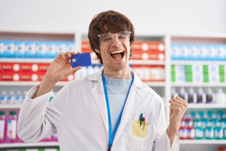 Téléchargez les photos : Jeune homme hispanique travaillant à la pharmacie pharmacie tenant carte de crédit criant fier, célébrant la victoire et le succès très excité avec bras levé - en image libre de droit