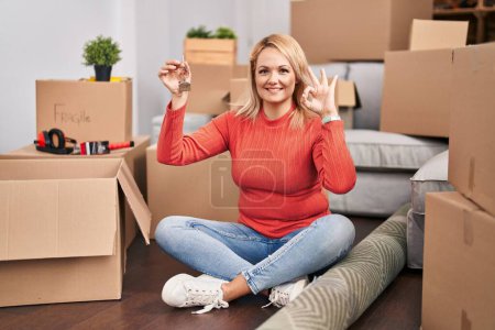 Photo for Blonde woman holding keys of new home sitting on the floor doing ok sign with fingers, smiling friendly gesturing excellent symbol - Royalty Free Image