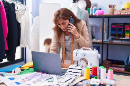 Téléchargez les photos : Middle age woman tailor talking on smartphone using laptop at clothing factory - en image libre de droit