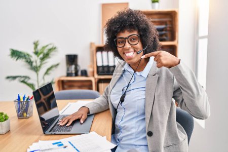 Téléchargez les photos : Black woman with curly hair wearing call center agent headset at the office smiling cheerful showing and pointing with fingers teeth and mouth. dental health concept. - en image libre de droit