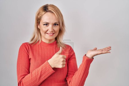 Foto de Mujer rubia de pie sobre un fondo aislado mostrando la palma de la mano y haciendo un gesto bien con los pulgares hacia arriba, sonriendo feliz y alegre - Imagen libre de derechos