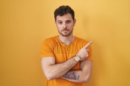 Téléchargez les photos : Jeune homme hispanique debout sur fond jaune pointant avec le doigt de la main sur le côté montrant la publicité, visage sérieux et calme - en image libre de droit
