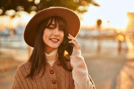 Mujer morena con sombrero de invierno sonriendo hablando por teléfono al aire libre en la ciudad al atardecer