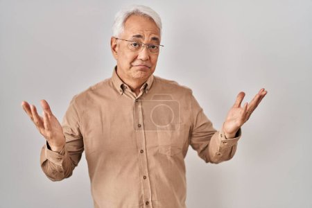 Foto de Hispanic senior man wearing glasses clueless and confused expression with arms and hands raised. doubt concept. - Imagen libre de derechos