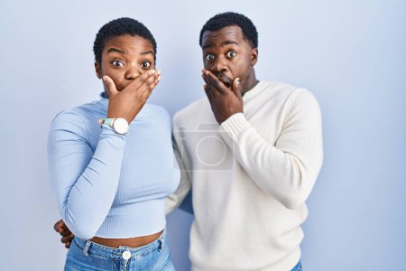 Foto de Joven pareja afroamericana de pie sobre fondo azul sorprendió cubriendo la boca con las manos por error. concepto secreto. - Imagen libre de derechos