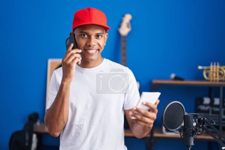 Photo for Young latin man musician talking on smartphone reading notebook at music studio - Royalty Free Image