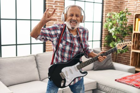 Foto de Senior hombre de pelo gris tocando la guitarra eléctrica haciendo cuernos símbolo con la mano en casa - Imagen libre de derechos