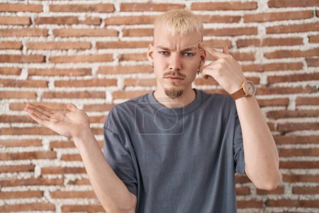 Young caucasian man standing over bricks wall confused and annoyed with open palm showing copy space and pointing finger to forehead. think about it. 