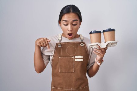Photo for Young hispanic woman wearing professional waitress apron holding coffee pointing down with fingers showing advertisement, surprised face and open mouth - Royalty Free Image