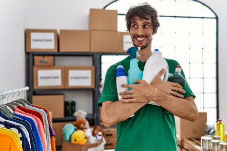 Photo for Young hispanic man wearing volunteer uniform holding cleaning products at charity center - Royalty Free Image