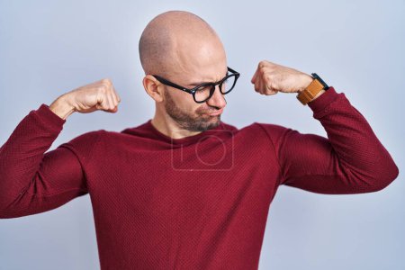 Téléchargez les photos : Jeune homme chauve avec la barbe debout sur fond blanc portant des lunettes montrant les muscles des bras souriant fier. concept de fitness. - en image libre de droit